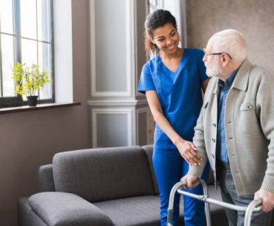 Portrait of an african young nurse helping old elderly disable man grandfather to walk using walker equipment in the bedroom. Senior patient of nursing home moving with walking frame and nurse support