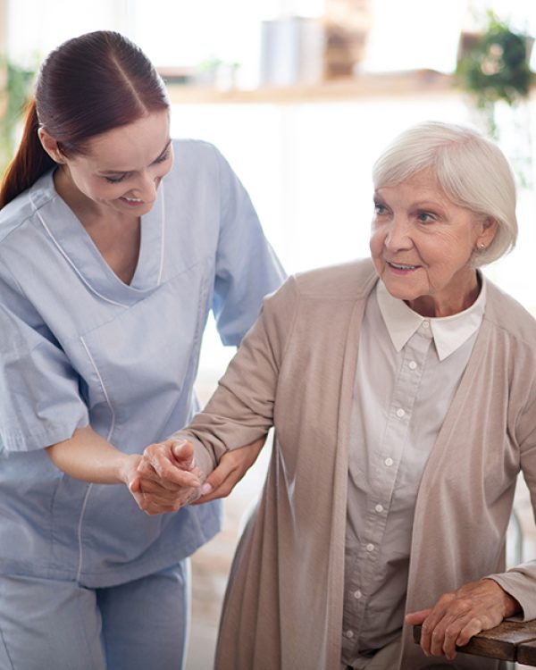 Helping to walk. Caregiver wearing uniform smiling while helping aged woman to walk