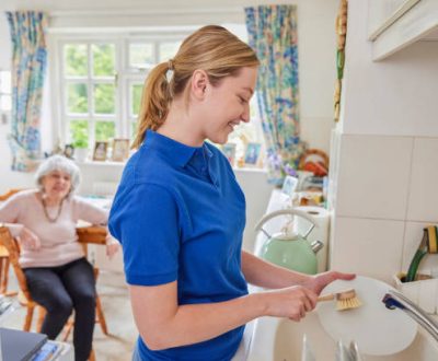 Female Home Help Cleaning House Doing Washing Up In Kitchen Whilst Chatting With Senior Woman